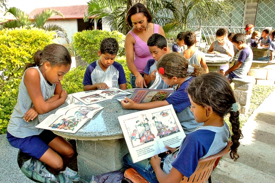 Crianças lendo livros infantis ao ar livre, acompanhados da professora