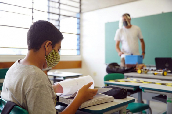 Criança, com máscara, sentada em carteira escolar, acompanha aula. Ao fundo, professor de máscare e face shield ao lado de quadro negro