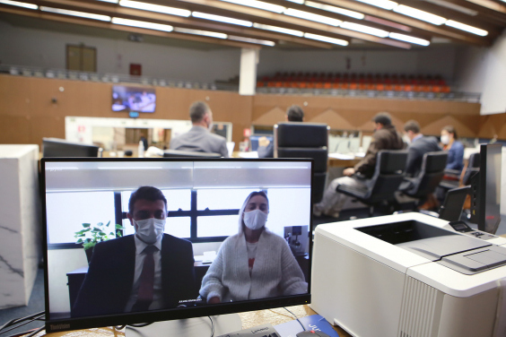 Monitor de computador exibe convidados em videoconferência. Ao fundo, vereadores compõem mesa de reunião no Plenário Amynthas de Barros. Galerias vazias mais ao fundo
