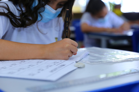 Menina sentada, inclinada sobre carteira escolar, com máscara no rosto, preenche um caderno educativo com lápis. Ao fundo, outro estudante realiza a mesma atividade,