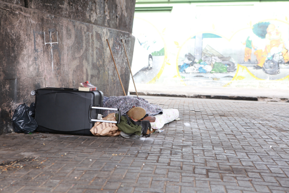 Homem com cabelos curtos pintados de amarelo está deitado no chão com um cobertor, ao lado de uma mala e debaixo de um viaduto . Ao fundo, um muro com pinturas urbanas que mostram lixo descartado irregularmente debaixo de um  viaduto