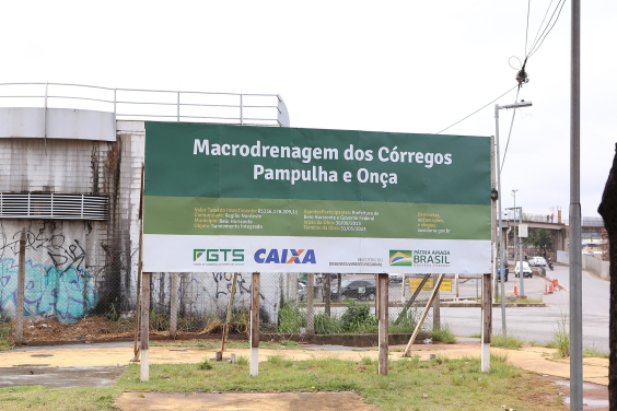 Placa indicativa de obras nos córregos Pampulha e Onça. Com fundo preto e letras brancas, a placa traz logomarcas do FGTS, da Caixa Econômica Federal, do Ministério de Desenvolvimento Regional e do Governo Federal