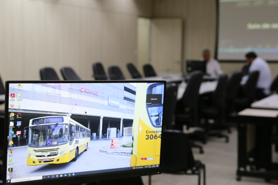 Imagem na tela do computador mostra ônibus da linha 328, amarelo, contornando um canteiro central em frente a um grande estabelecimento comercial . ao fundo, imagem desfocada de dois vereadores sentados à mesa
