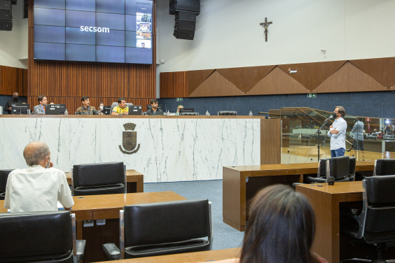 Quatro homens sentados à mesa do Plenário Amynthas de Barros e um homem falando ao microfone, à direita. Ao fundo, quatro assessores da Câmara.