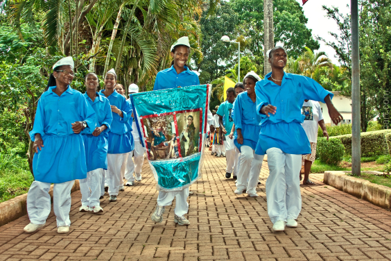 Homens e mulheres negros vestidos de azul e branco levam um estandarte  com imagem de santo por uma alameda arborizada
