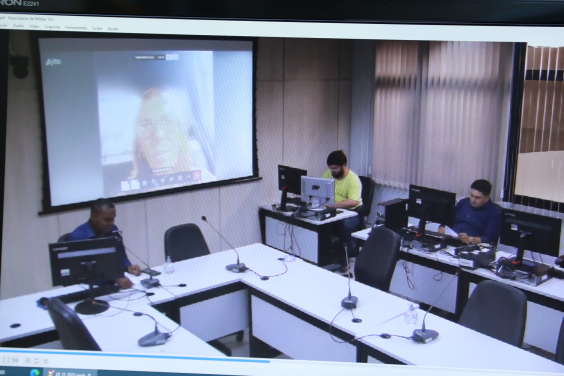 Sentado à mesa, em frente a computador, vereador acompanha reunião remota. À sua direita, dois assessores da Câmara, sentados em mesas individuais e em frente e computadores, o acompanham.