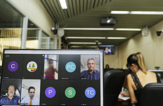 Quatro parlamentares dividem a tela de computador em reunião remota. Ao fundo, Plenário Camil Caram, com assessora da Câmara sentada, acompanhando. 