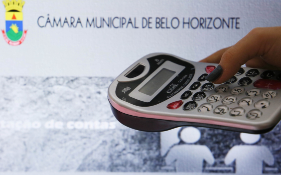 Foto mostra mão segurando calculadora com fundo sobre imagem ilustrativa da CMBHãmara 