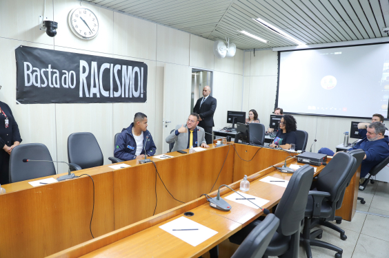 Imagem dos participantes da Audiência Pública. Uma faixa negra pendurada na parede traz os dizeres: Basta ao racismo