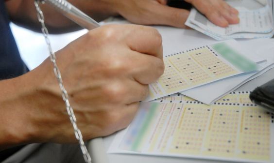 Mãos humanas preenchem boletos de loteria com números em sequência, em cima de um balcão. 