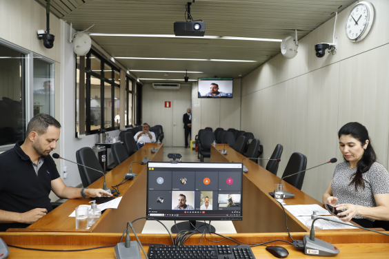 dois parlamentares sentados à mesa, em reunião presencial, e dois participando remotamente, exibidos em tela de computador.ticipando 