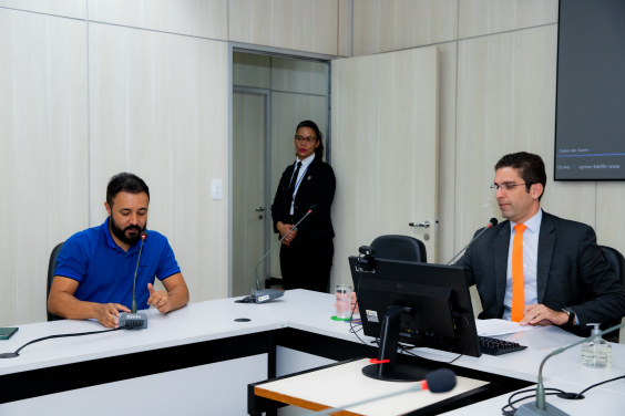 Os vereadores Braulio Lara e José Ferreira estão sentados no plenário. Uma segurança está em pé ao fundo, junto à porta.