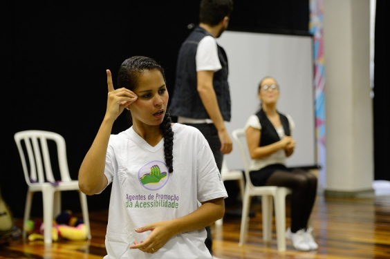 Mulher com camisa branca escrito "Agentes de promoção da acessbilidade", faz um sinal em Libras 