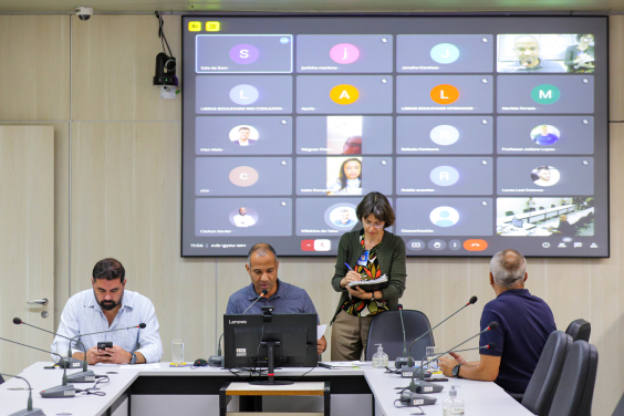 Vereadores estão sentados no plenário Helvécio Arantes. O telão atrás deles mostra a videochamada com parlamentares presentes virtualmente.