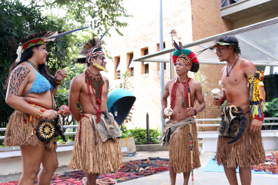 Pessoas indígenas tocam instrumentos de percussão em vestes tradicionais na frente da entrada da CMBH