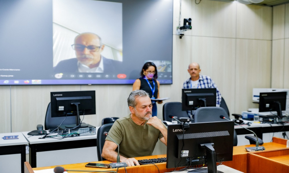 Vereador Wilsinho da Tabu sentado em frente ao computado, com a tela atrás projetando a imagem do vereador Fernando Luiz.