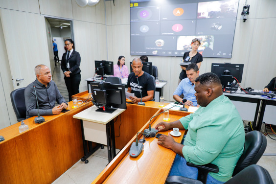 Vereadores assentados no Plenário Camil Caram em reunião da comissão, telão atrás deles mostra vereadores presentes em chamada virtual