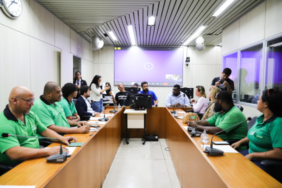 Vereadores e convidados da audiência estão sentados no plenário. O telão atrás deles mostra a videochamada com participantes presentes virtualmente.