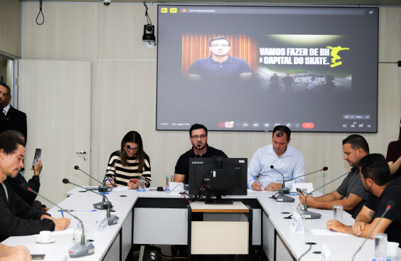 Vereadores e convidados assentados no Plenário Helvécio Arantes durante audiência pública. O telão atrás deles mostra apresentação.