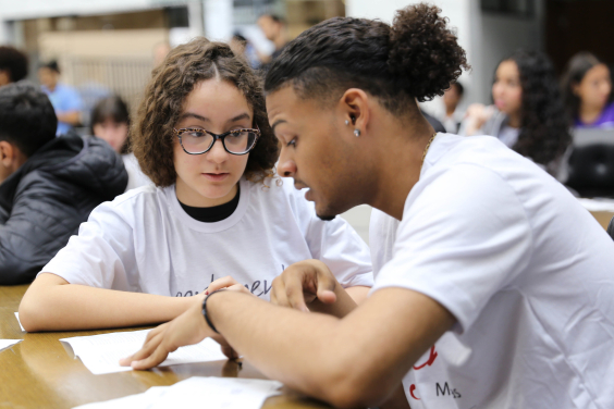 Imagem de dois jovens, um negro e um caucasiano, ambos usando camiseta branca sentados à mesa conversando