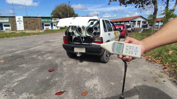 carro com som automotivo instalado no porta malas; close nas mãos de pessoa com instrumento de aferição de ruído