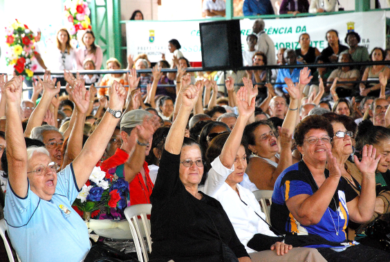 pessoas idosas assentadas levantam os braços participando de evento