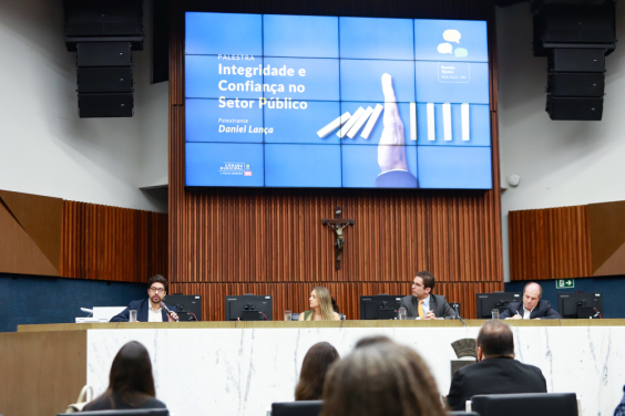 Mesa do Plenário Amintas de Barros coma  presença do vereador Braulio Lara e outros participantes. No telão, está o escrito "Palestra Integridade e Confiança no Setor Público, palestrante Daniel Lança".