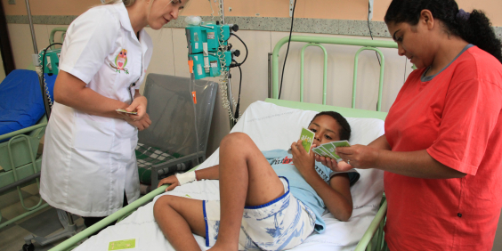 Criança em leito hospitalar acompanhada de mulher e atendida por profissional de saúde.