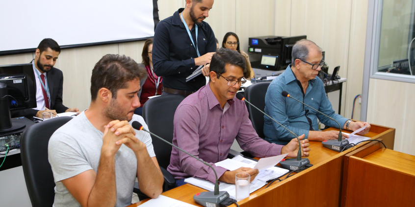 Três integrantes da comissão estão sentados à Mesa do Plenário Camil Caram; um servidor de pé e três assentados atrás deles assessoram e registram as ocorrências da reunião.