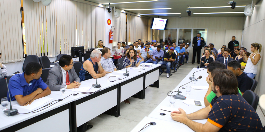 Vista aberta do plenário. Parlamentares e convidados compõem mesa de reunião. Ao fundo, pessoas acompanham a reunião.