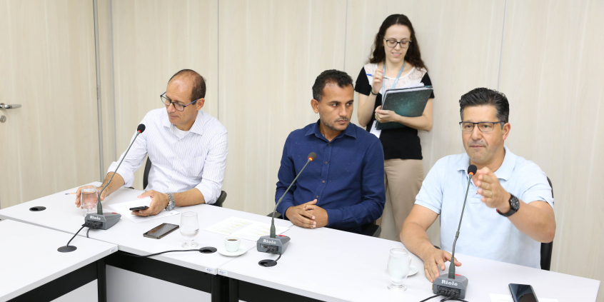 Integrantes da Comissão estão sentados à Mesa apreciando a pauta da reunião; atrás, de pé, uma assessora segura uma pasta de papéis