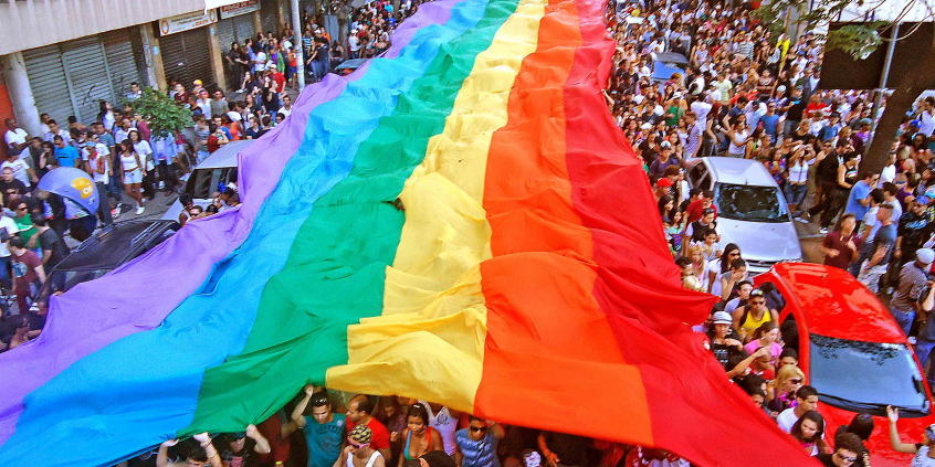 manifestação popular em via pública; pessoas erguem bandeirão com o arco-íris