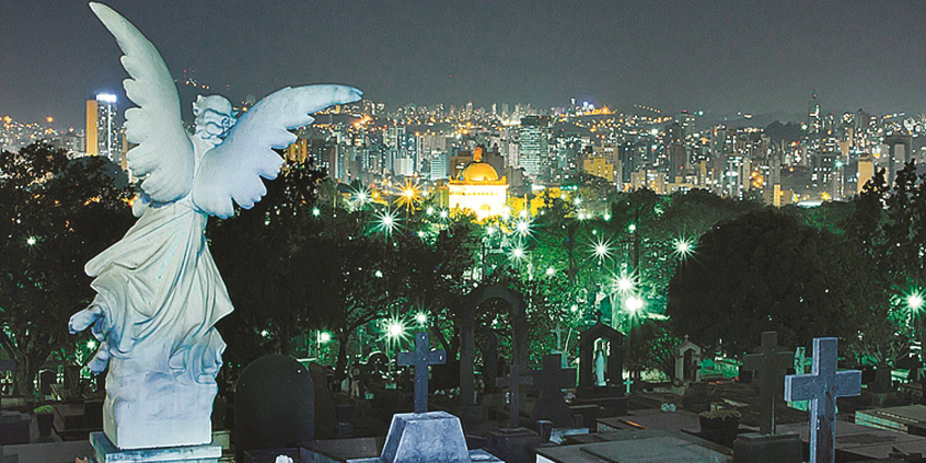 Vista noturna de lápides e esculturas no Cemitério do Bonfim. Ao fundo, luzes da cidade. Vista panorâmica da cidade