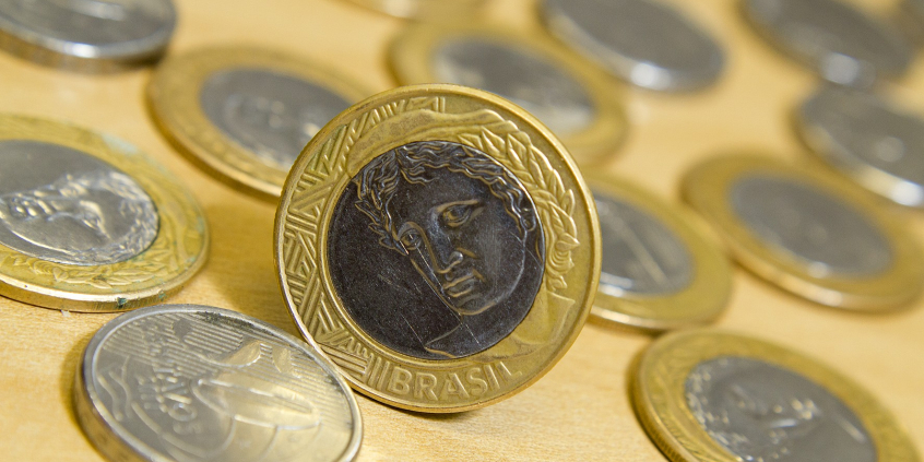 moedas de real deitadas sobre a mesa. Uma delas em destaque, em pé