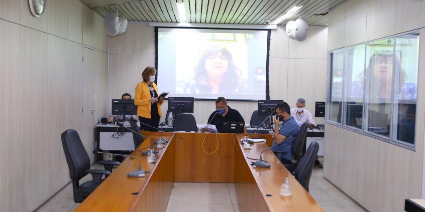 Vereadores compõem mesa de reunião. Telão ao fundo em videoconferência