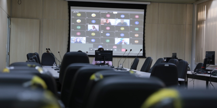 imagem de monitor, ao fundo. Em primeiro plano, vista aberta do Plenário, com cadeiras vazias. O monitor exibe vereadores em videoconferência