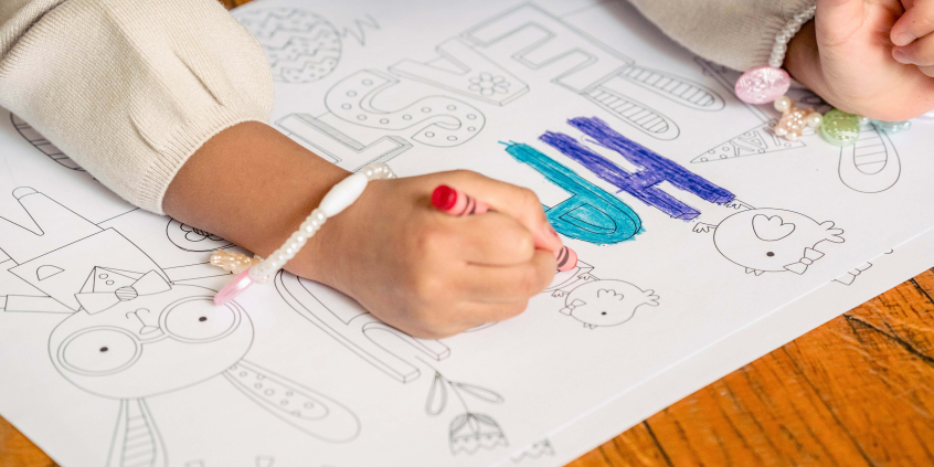 Mãos de criança sobre cartaz ilustrado, colorindo desenhos. Cartaz sobre piso de madeira