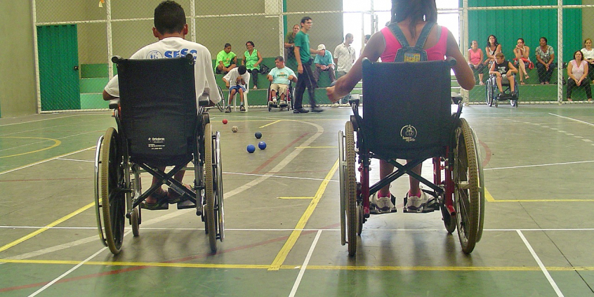 Dois estudantes cadeirantes, de costas, em quadra esportiva, durante o dia; no meio, cinco bolas pequenas no chão. Ao fundo, três estudantes cadeirantes e um homem de pé. Mais ao fundo, atrás de uma grade de proteção, cerca de dez adultos acompanham sentandos em arquibancada. 