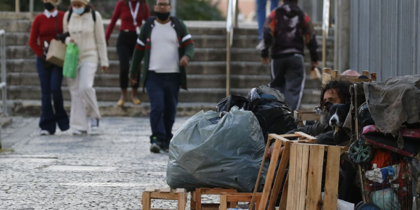 Morador de rua cercado de pertences, sentado em rua de cidade encostado em muro, com seis cidadãos passando ao fundo, próximo a uma escada de seis degraus, durante o dia.