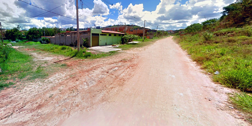 Rua sem asfalto com casas sem reboco à esquerda, durante o dia.