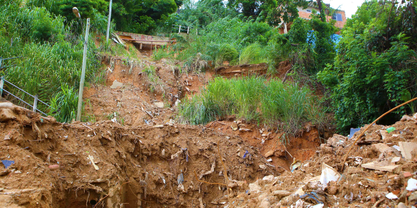 Barranco com terra e vegetação nas laterais; no alto, ao fundo, uma residência. Imagem diurna.