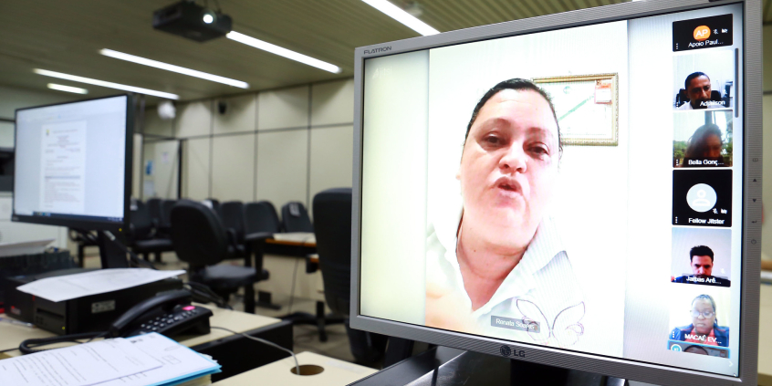 Mulher fala, em destaque, em reunião remota, na tela de um computador. Do lado direito da tela estão seis outras janelas menores, com imagens de dois homens, duas mulheres e duassomente com iniciais de nomes.