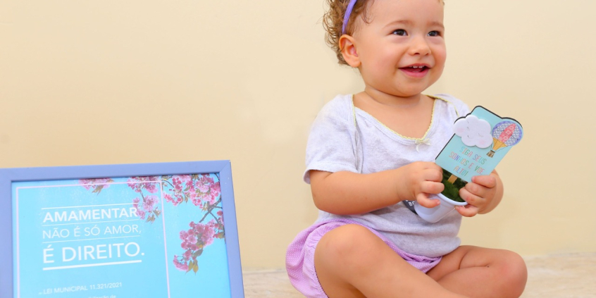 Criança caucasiana usa lado de fita lilás no cabelo, blusa branca e short lilás. ao lado, um quadro com os dizeres: "Amamentar não é só amor. É um direito."