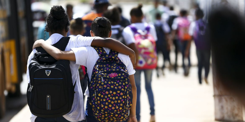 Duas estudantes, de costas, cada uma com uma mochila, abraçadas, caminham, durante o dia. Ao fundo, mais estudantes.