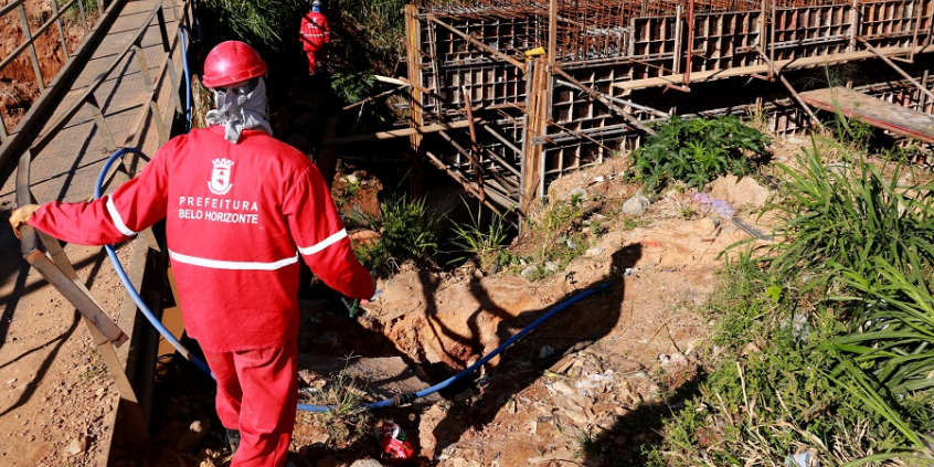 Dois funcionários de obras com uniformes e capacetes vermelhos andar por obra de grande porte da Prefeitura.