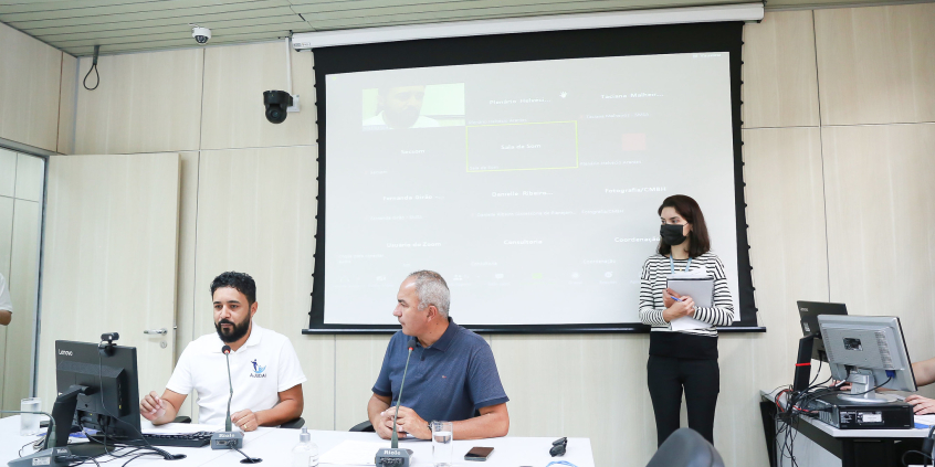 Os vereadores José Ferreira e Claudio do Mundo Novo estão sentado à mesa de reunião. Ao fundo e no alto, o telão mostra a imagem dos outros participantes da reunião. Em pé, uma mulher usando uma blusa listrada, calça preta e máscara, segura uma pasta de documentos