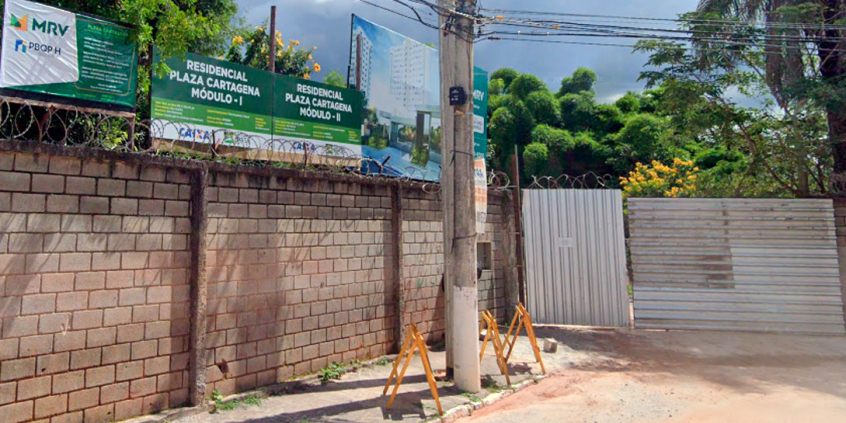 Muro de construção com Cartaz de duas torres de prédios com mais de dez andares ao fundo e dois cartazes indicando Residenciais Cartagena, Módulos I e II, à frente, durante o dia.