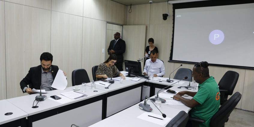 Quatro vereadores sentados à mesa, em reunião presencial, com dois assessores da Câmra ao fundo.