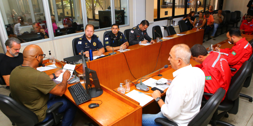 três vereadores reunidos com representantes da Guarda Civil Municipal, do Corpo de Bombeiros Militar de Minas Gerais e dos bombeiros civis, sentados à mesa. Ao fundo, Brigadistas e bombeiros assistem à reunião, sentados.