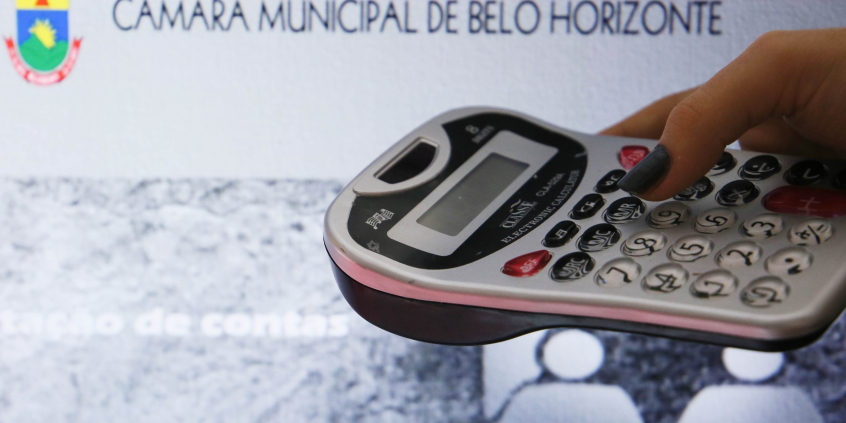 Foto mostra mão segurando calculadora com fundo sobre imagem ilustrativa da CMBHãmara 